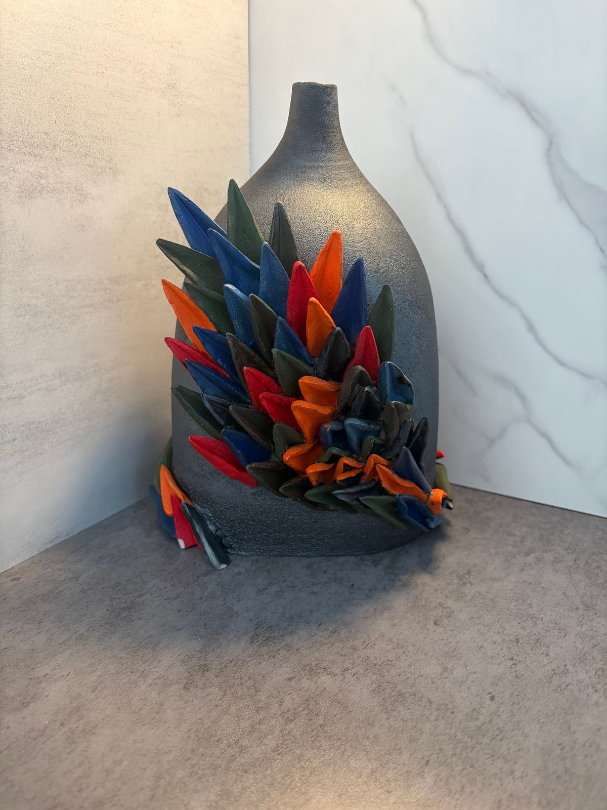 Front-facing view of handmade ceramic totem with dense modular glazed spikes in cobalt, orange, red, and forest green applied across the lower half of a matte dark vessel