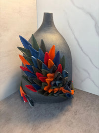 Dark stoneware ceramic vase with colorful sculptural leaf projections in blue, green, red, and orange, photographed from a three-quarter angle against marble and concrete surfaces